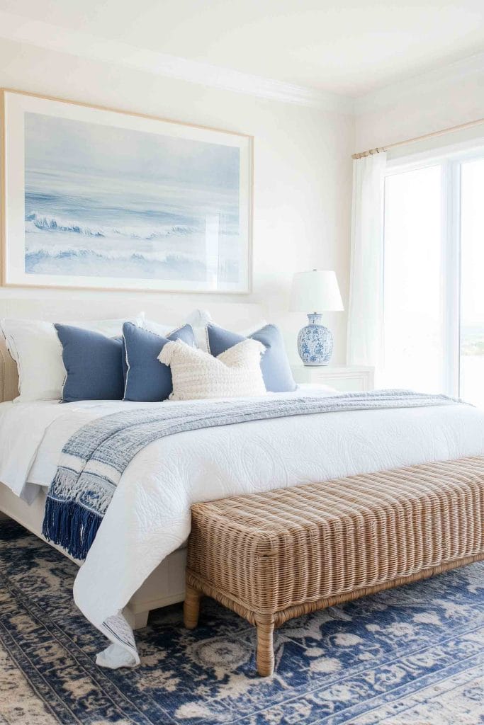 A classic coastal bedroom with a white bed frame and blue accents. The bed is styled with a mix of blue and white pillows, and a white quilted coverlet with a blue tasseled throw. A large seascape painting hangs above the bed, and a wicker bench rests at the foot of the bed. Natural light pours in from the large windows, and a blue and white ceramic table lamp adds a coastal touch.