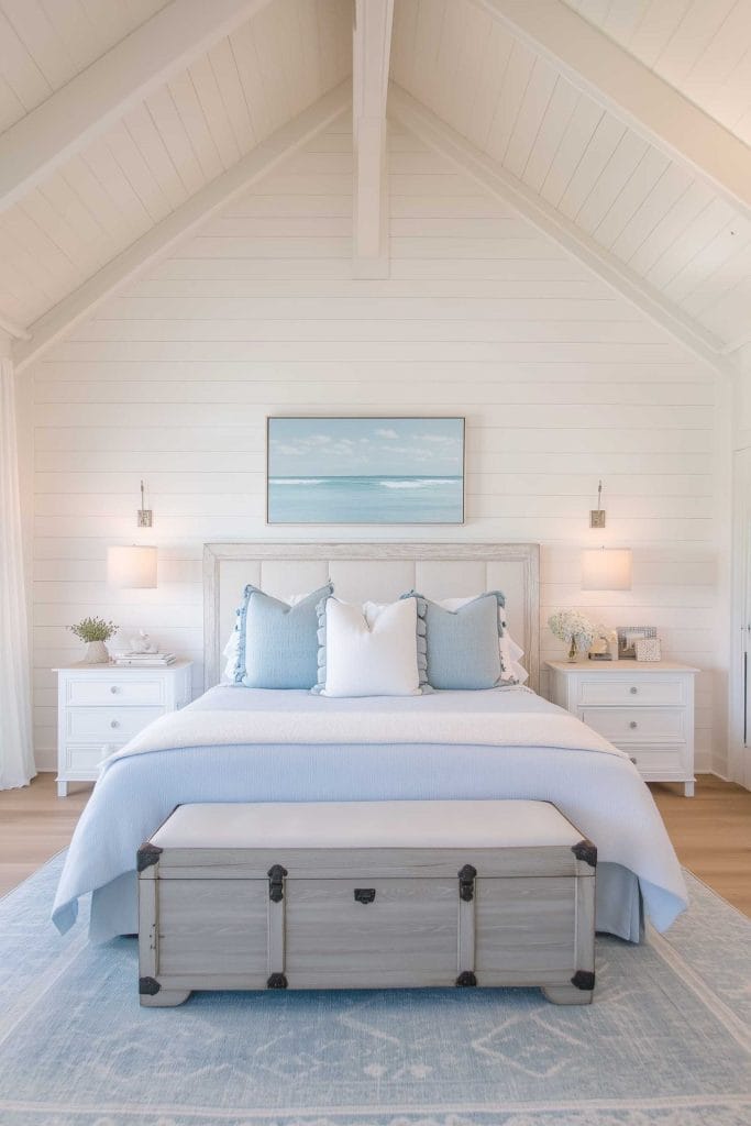 This cozy bedroom has a vaulted ceiling and features a simple white wooden bed frame. The bedding consists of white linens with pale blue pillows. A framed ocean painting hangs on the white shiplap wall, with matching bedside tables and lamps on each side. A light blue rug under the bed and a gray chest at the foot create a beachy yet elegant aesthetic.