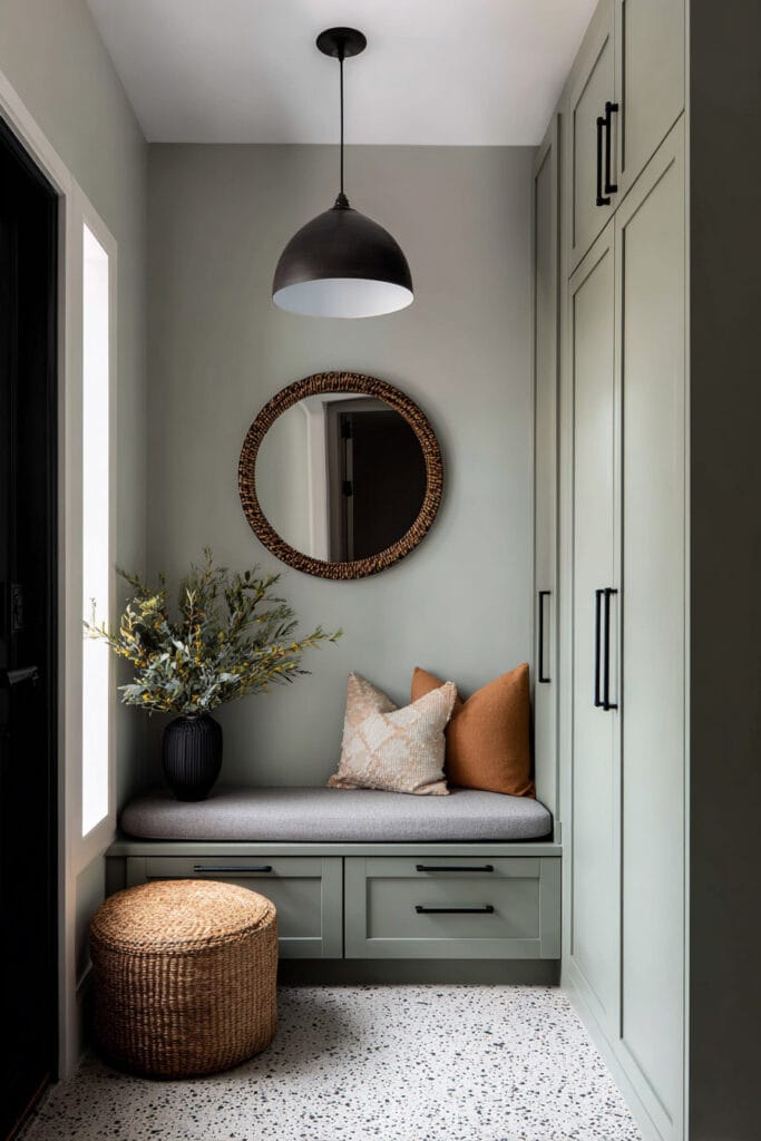 Modern sage green entryway with built-in cabinets, bench seating, round woven mirror, black pendant light, and cozy pillows.
