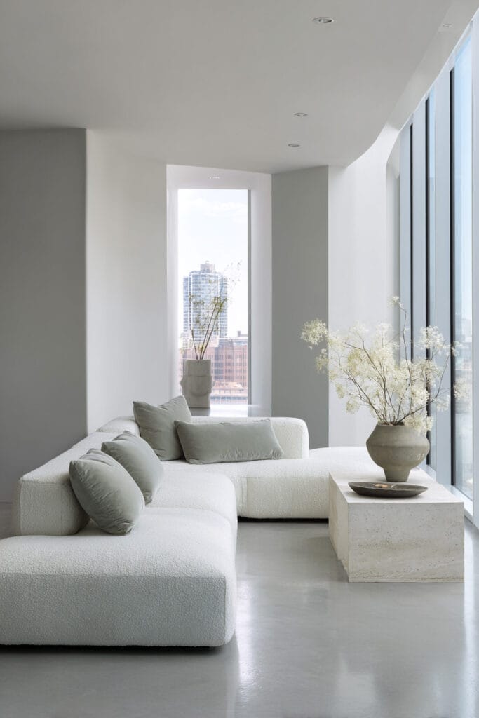 Modern living room with white boucle sofa, sage green pillows, floor-to-ceiling windows, and minimalist stone decor.