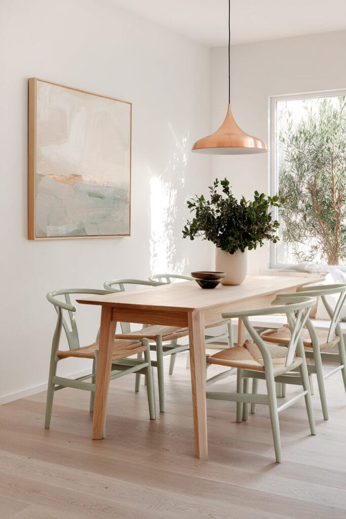 Dining room with sage green wishbone chairs, natural wood table, copper pendant light, greenery, and abstract wall art.