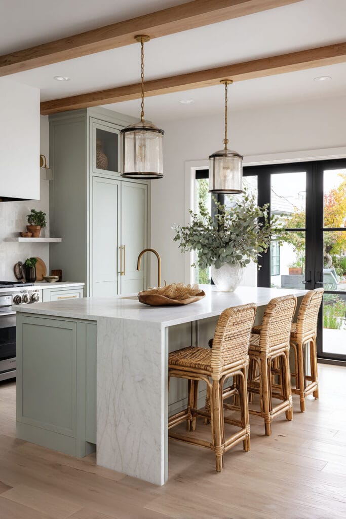 Sage green kitchen with marble waterfall island, woven barstools, brass fixtures, and wood ceiling beams.