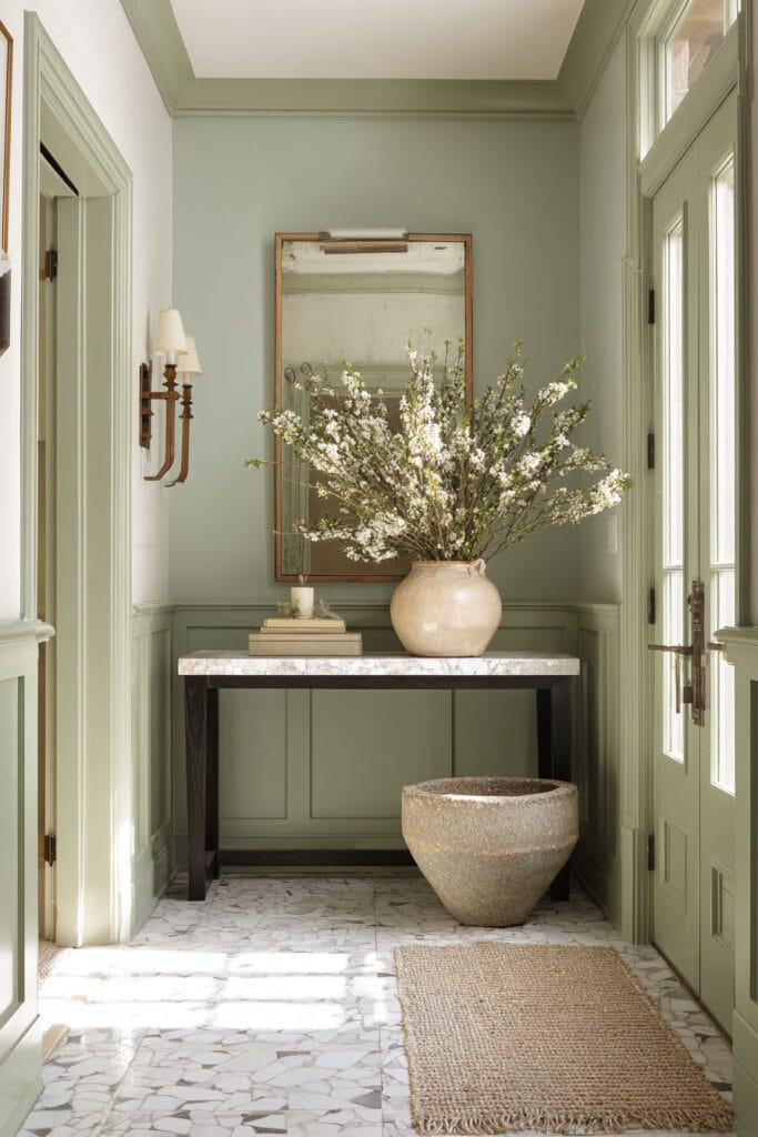 Sage green entryway with marble console table, ceramic vase of blooming branches, woven rug, and vintage wall sconces.