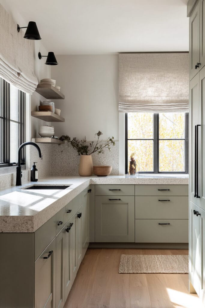Sage green kitchen with terrazzo countertops, black hardware, floating wood shelves, and natural light from Roman shades.