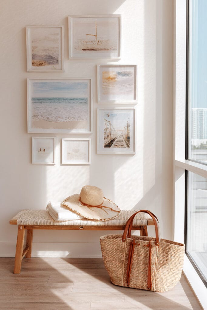 Coastal gallery wall with beach and ocean prints in white frames, woven bench, straw hat, and beach bag in sunlit corner decor.
