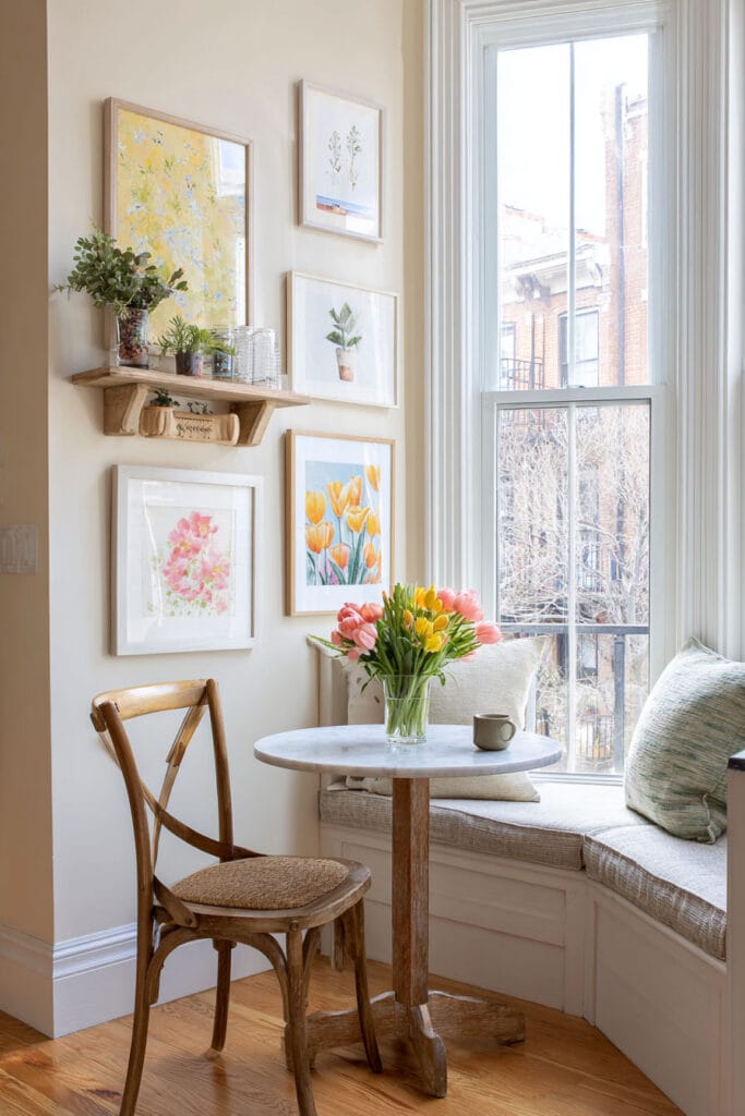 Sunny breakfast nook with floral gallery wall, marble bistro table, tulips, and layered wall shelf with plants.