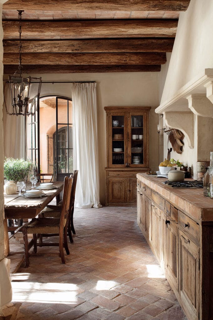 Rustic Mediterranean kitchen with terracotta tile floors, reclaimed wood cabinets, exposed beams, plaster walls, and soft white curtains. Warm earthy palette with European farmhouse charm.