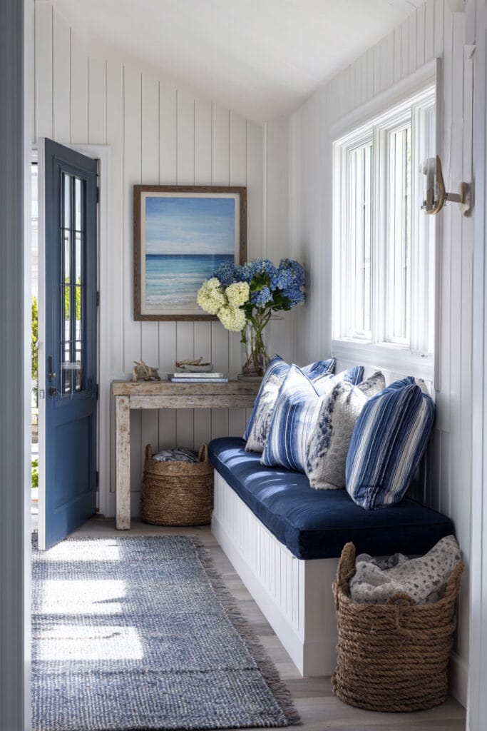 Coastal entryway with navy blue bench cushions, striped pillows, woven baskets, white shiplap walls, and ocean artwork. Fresh beach-inspired blue and white color palette.