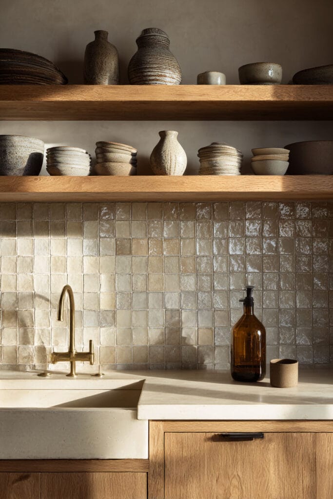 Warm neutral kitchen with rustic wood shelves, handmade ceramic dishes, zellige tile backsplash, natural oak cabinets, brass faucet, and stone countertop.