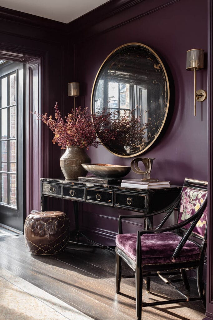 Aubergine room with black console, round antique mirror, velvet chair, and brass sconces.