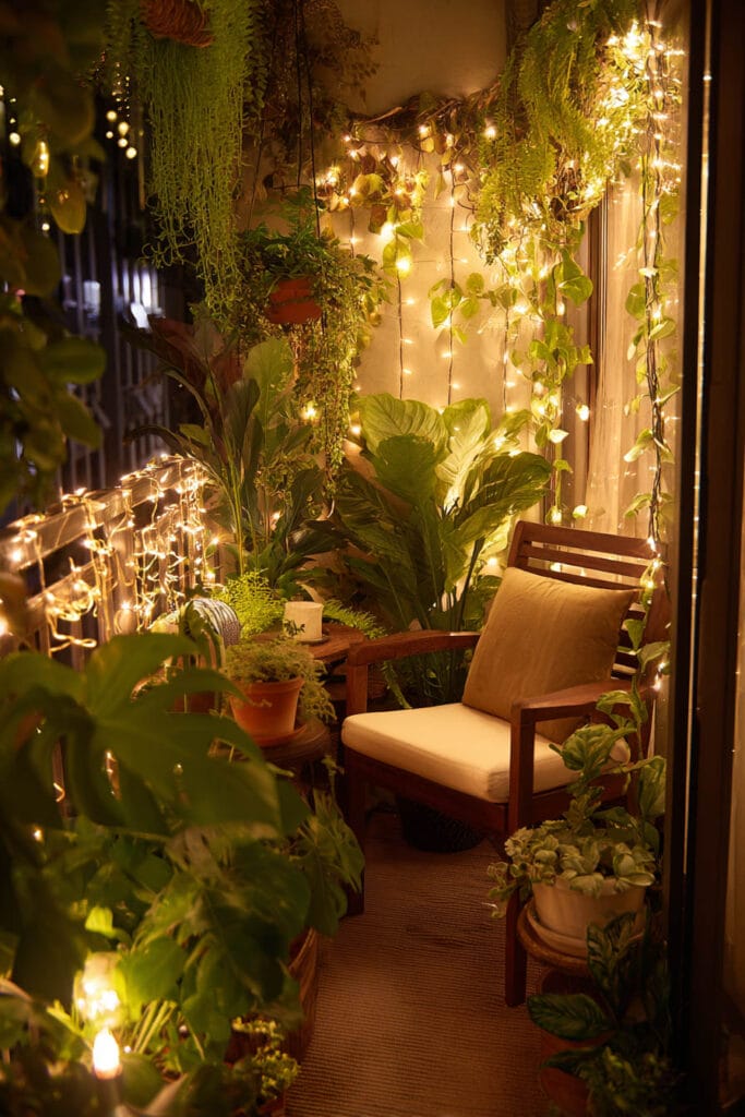 Modern balcony with warm string lights, lush tropical plants, wooden lounge chair, and ambient lighting creating an elevated luxury outdoor retreat at night.