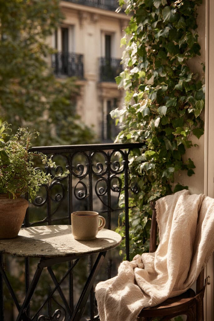 Small Paris balcony with coffee cup, wrought iron railing, climbing ivy, cozy chair with throw blanket, and morning sunlight
