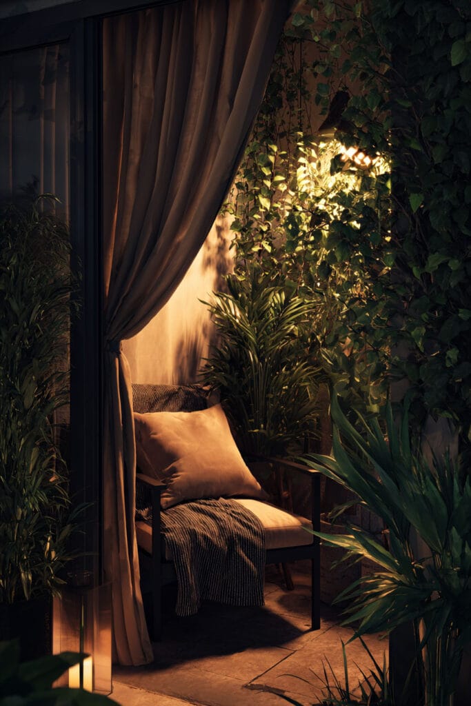 Private balcony with warm wall lantern, curtains, lush greenery, cozy chair, and layered outdoor lighting for a relaxing nighttime retreat.