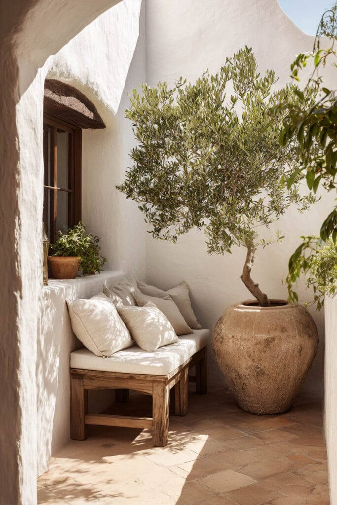 Mediterranean balcony with olive tree in oversized terracotta pot, rustic wood bench, neutral cushions, white stucco walls, and warm natural light.