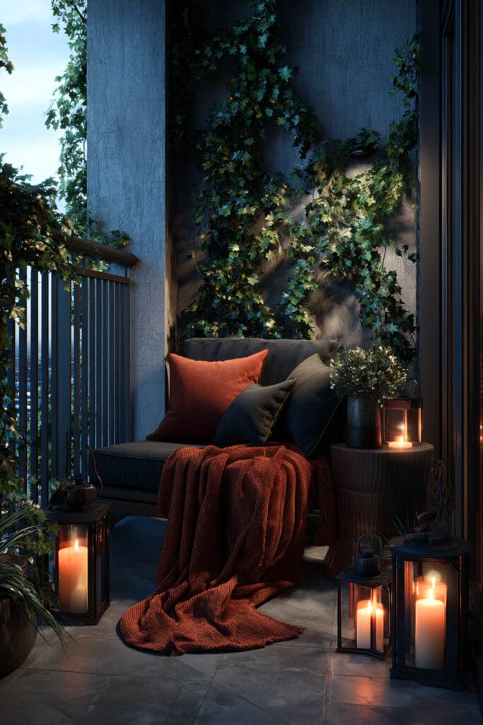 Moody balcony with candle lanterns, climbing vines, cozy seating, rust-colored throw blanket, dark walls, and warm ambient lighting.