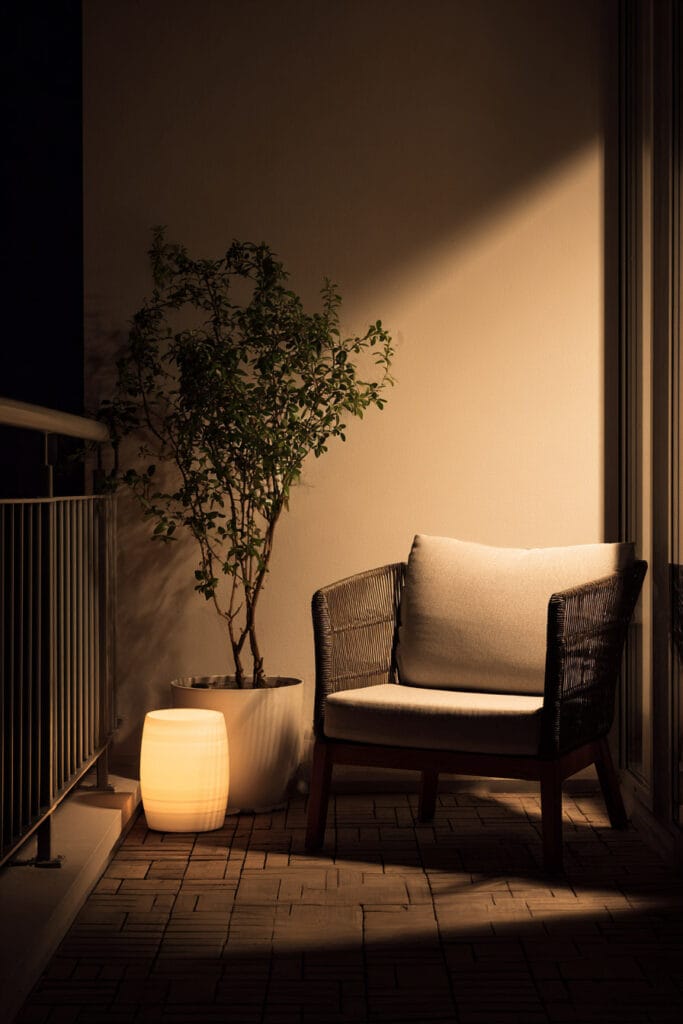 Modern balcony with glowing floor lamp, contemporary outdoor chair, potted tree, and warm ambient lighting creating a luxury minimalist retreat.