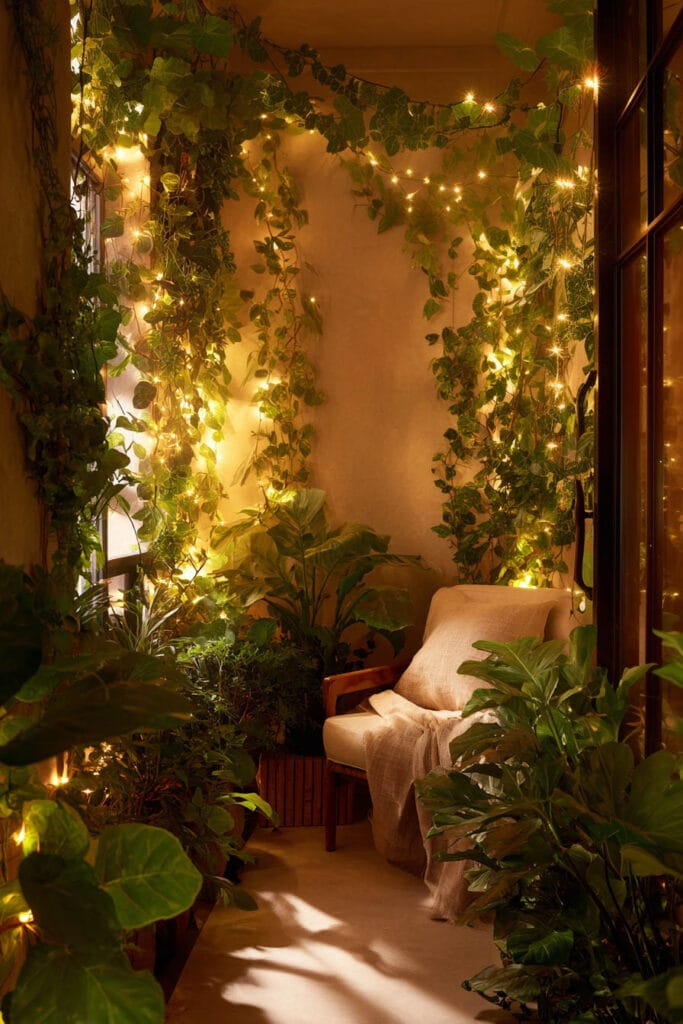 Cozy balcony with fairy lights woven through cascading vines, tropical plants, and comfortable seating creating a magical garden atmosphere at night.