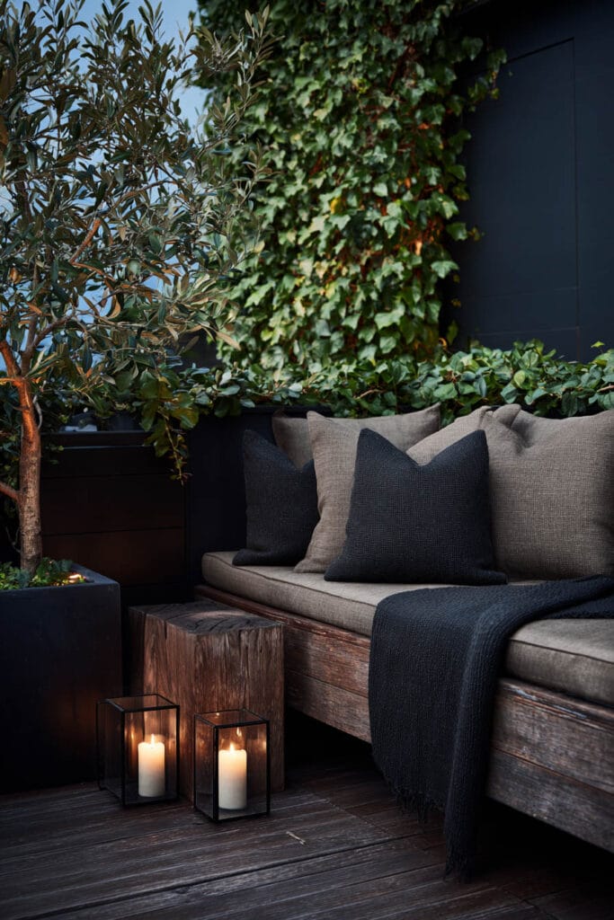 Moody balcony with olive tree, climbing ivy, dark throw pillows, candle lanterns, wooden bench seating, and layered greenery.