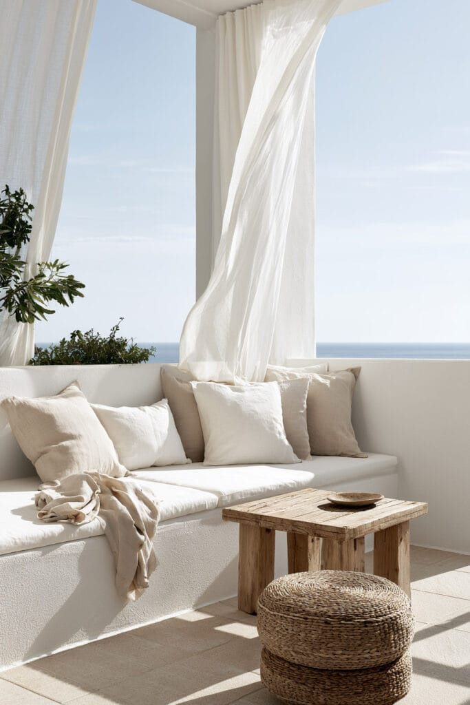 Cozy Greek island-inspired balcony with built-in white seating, flowing linen curtains, rustic wood table, woven ottoman, and ocean views in a bright European coastal setting.