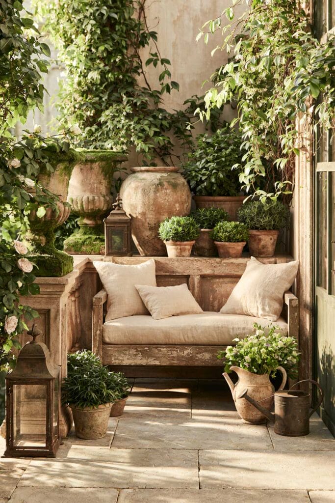 Hidden secret garden balcony with a rustic wooden loveseat, weathered stone planters, climbing vines, terracotta pots, and vintage European garden decor.