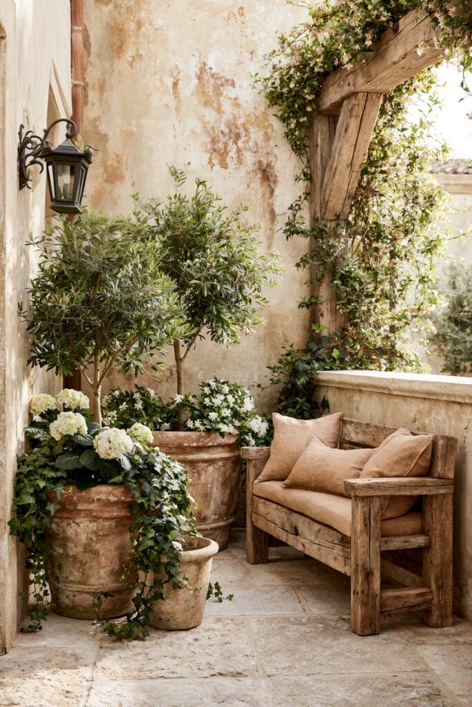 Secret garden balcony with rustic wooden bench, olive trees, terracotta pots, climbing jasmine, and European courtyard decor.