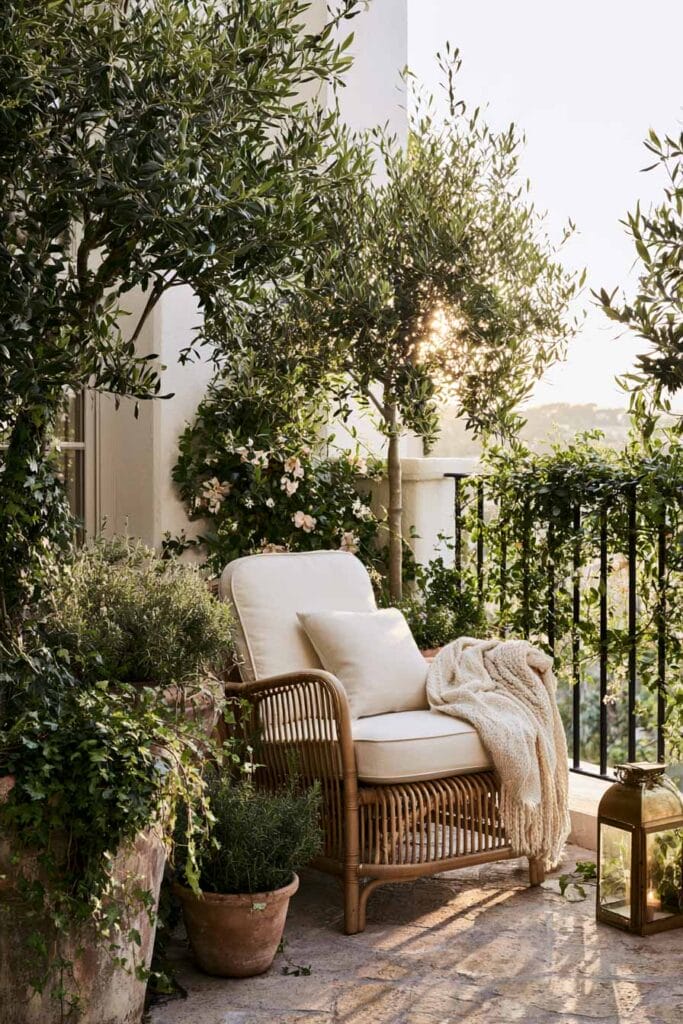 Romantic European balcony with a wicker lounge chair, olive trees, climbing vines, terracotta planters, lantern, and lush greenery creating a cozy hidden garden outdoor retreat.