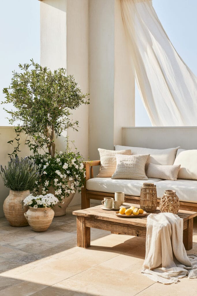 Beautiful European balcony with Mediterranean coastal decor, white stucco walls, olive tree, rustic wood coffee table, linen curtains, oversized pottery, and outdoor lounge seating.