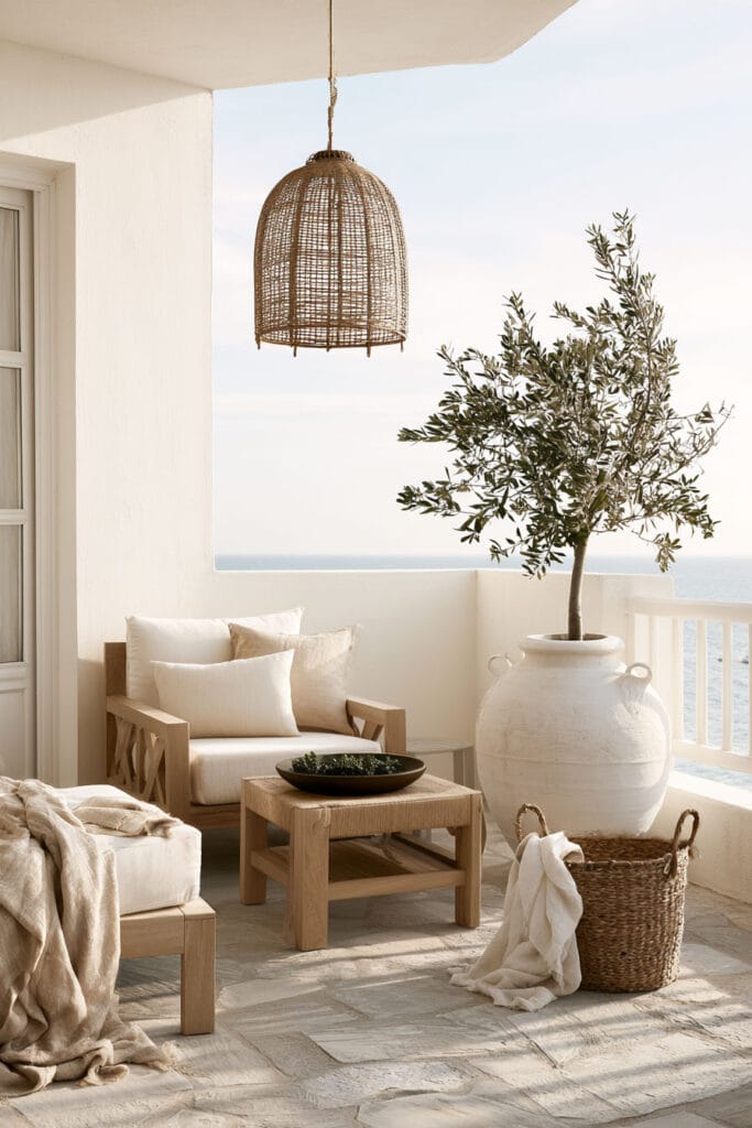 Cozy Mediterranean balcony with linen lounge chair, light wood furniture, oversized olive tree, woven pendant light, and ocean view in a bright European coastal setting.