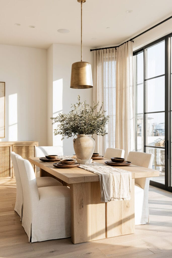 Bright neutral dining room with natural wood table, cream linen chairs, woven cane, pottery, olive branches, and dark brown accents.