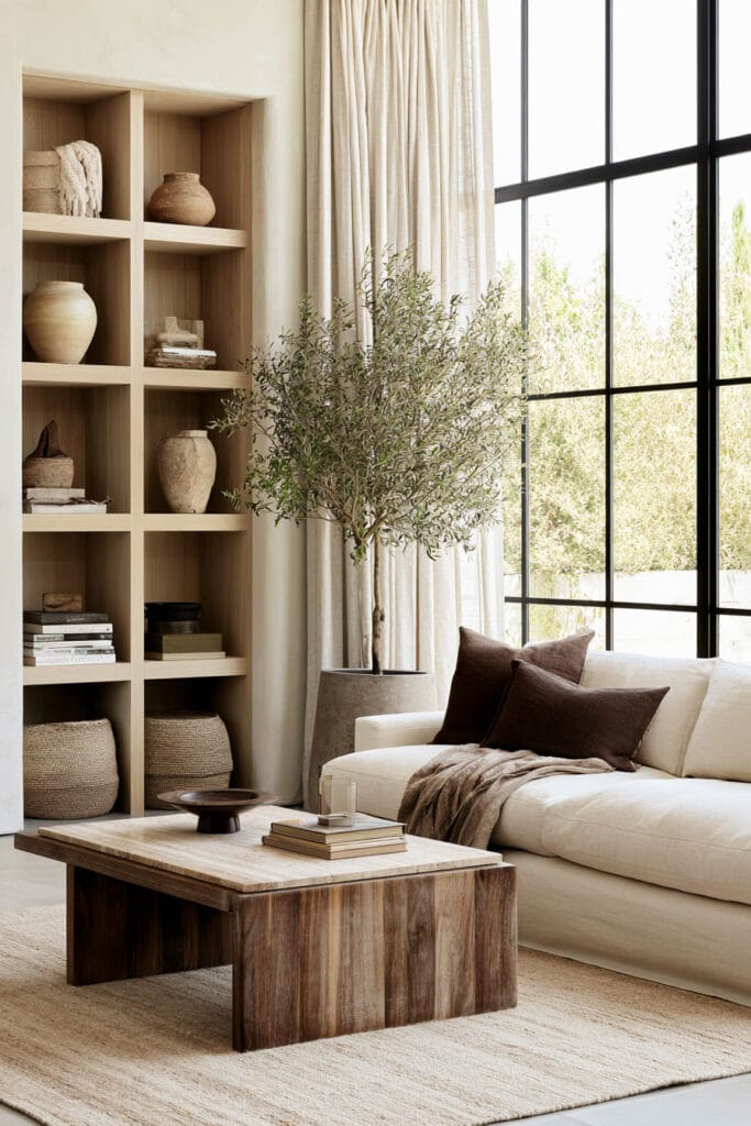 Cream sofa with chocolate brown pillows, natural wood coffee table, woven baskets, olive tree, and warm neutral home decor.