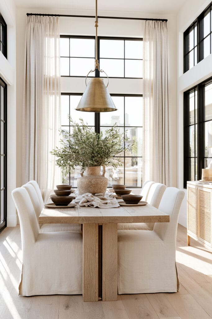 Warm neutral dining room with chocolate brown tableware, natural wood dining table, cream chairs, woven cane cabinet, and brass pendant.