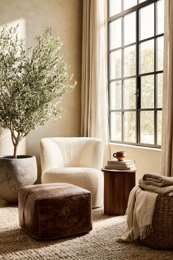 Bright reading corner with cream bouclé chair, chocolate brown leather ottoman, dark wood side table, olive tree, and warm neutral decor.