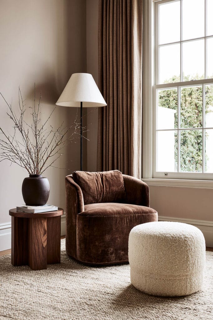 Warm minimalist reading nook with a chocolate brown velvet chair, dark wood side table, brown curtains, ivory bouclé ottoman, and neutral woven rug.