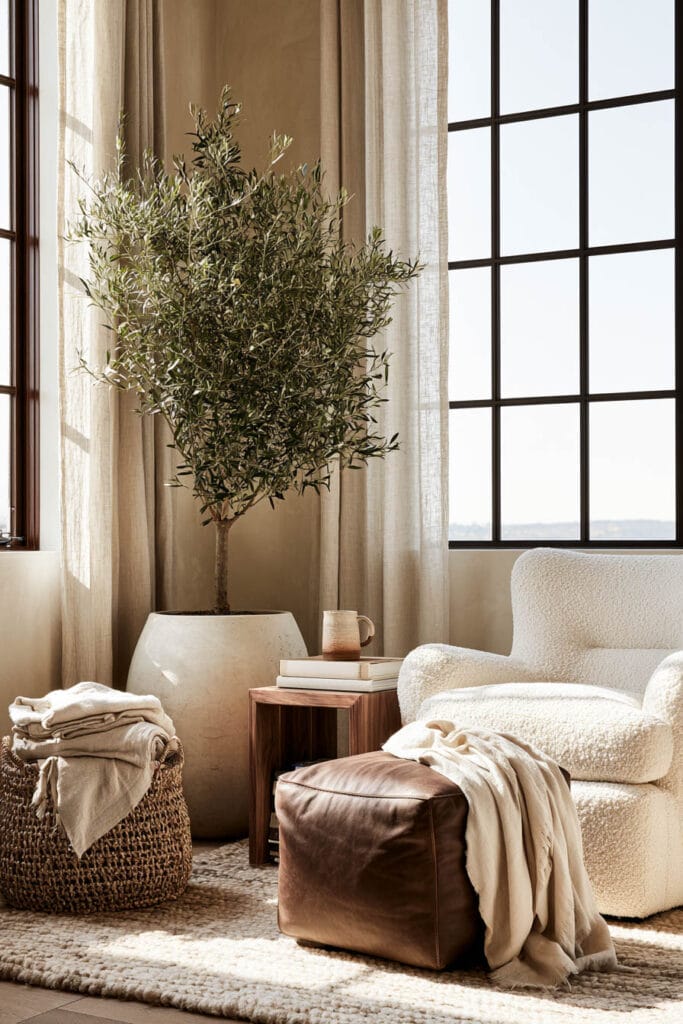Cream boucle chair with chocolate brown leather ottoman, dark wood side table, olive tree, woven basket, and warm neutral decor.