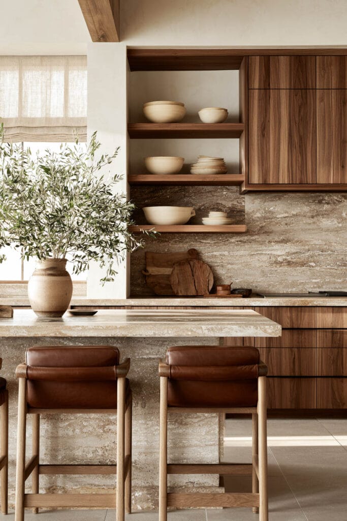 Chocolate brown wood kitchen cabinets with cognac leather stools, natural stone island, open shelving, cream ceramics, and organic decor.