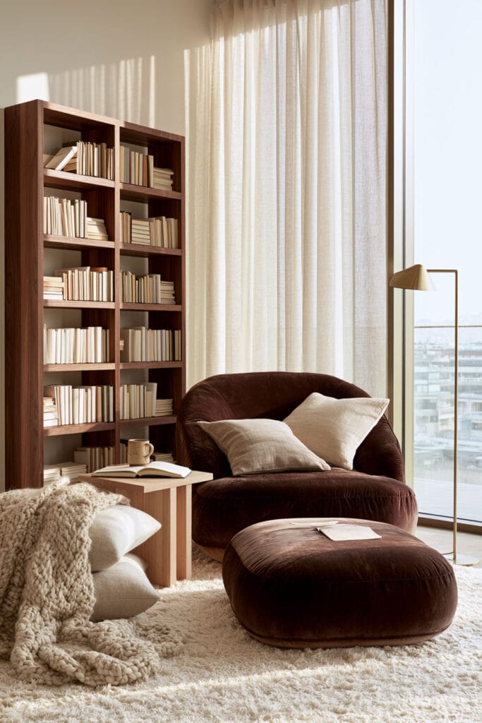 Chocolate brown velvet chair and ottoman with cream pillows, chunky knit throw, ivory rug, warm wood bookcase, and layered neutral textures.