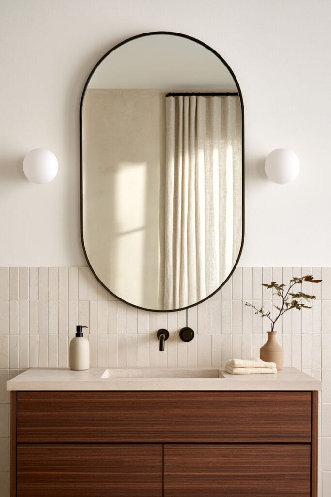 Tall arched bathroom mirror above a dark wood vanity with globe sconces, cream vertical tile, and warm neutral decor.