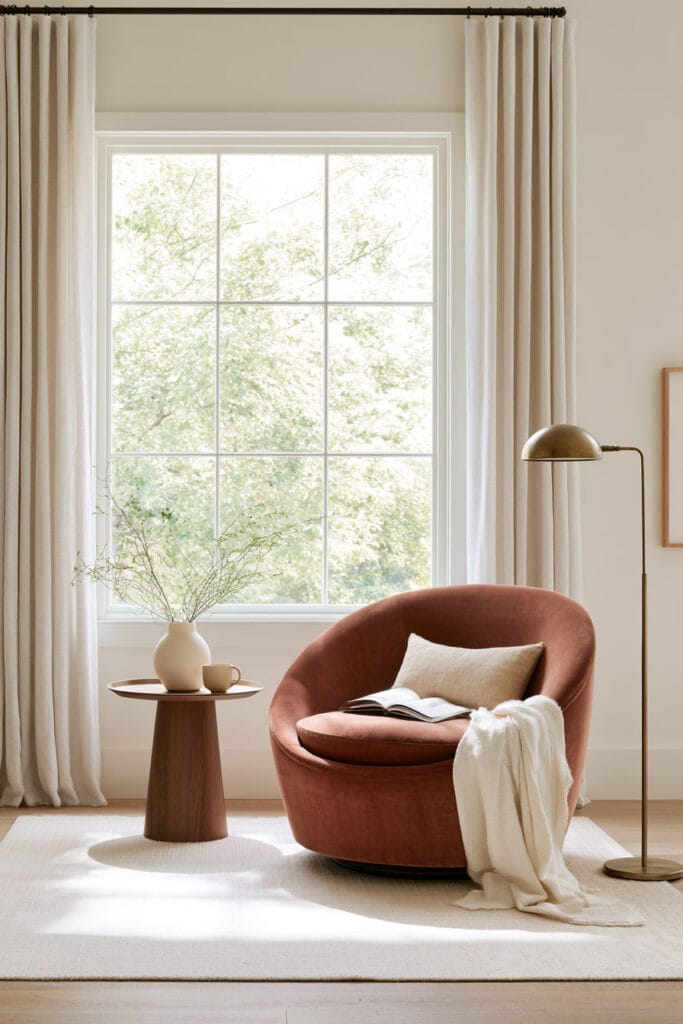 Rounded rust accent chair in a bright reading nook with round wood side table, brass floor lamp, cream curtains, and neutral rug.