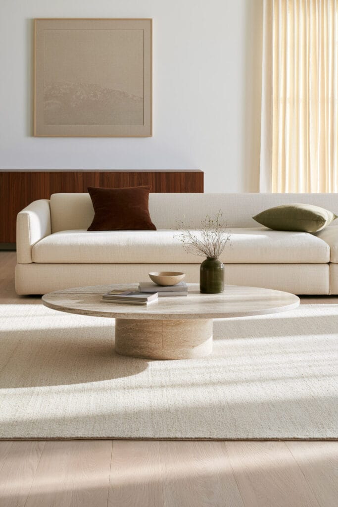 Round stone coffee table in a warm neutral living room with cream sofa, wood cabinetry, and earthy brown and olive accents.