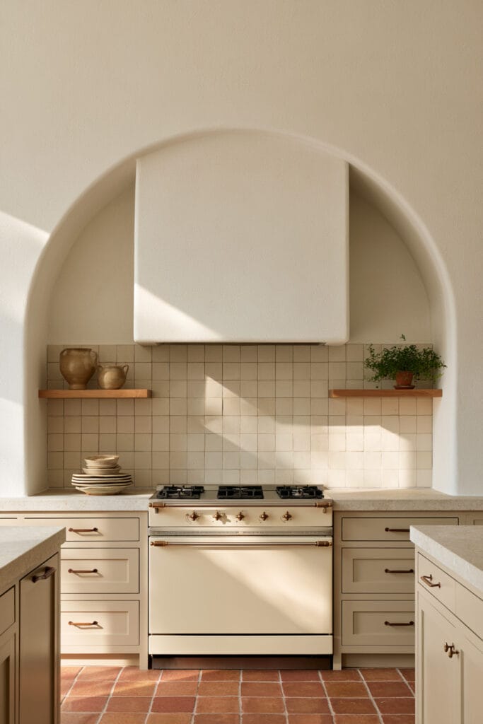 Warm neutral kitchen with large arched range alcove, cream hood, tiled backsplash, wood shelves, and terracotta flooring.