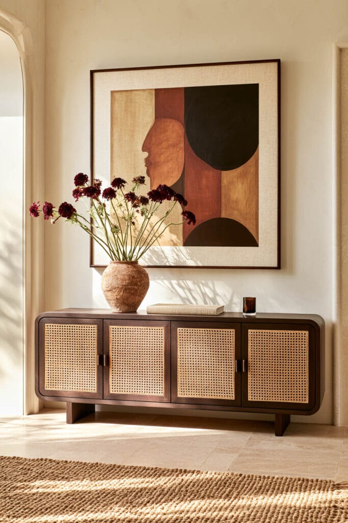 Afrohemian entryway with dark wood and cane credenza, earth-toned wall art, rustic pottery vase, burgundy flowers, and natural woven rug.