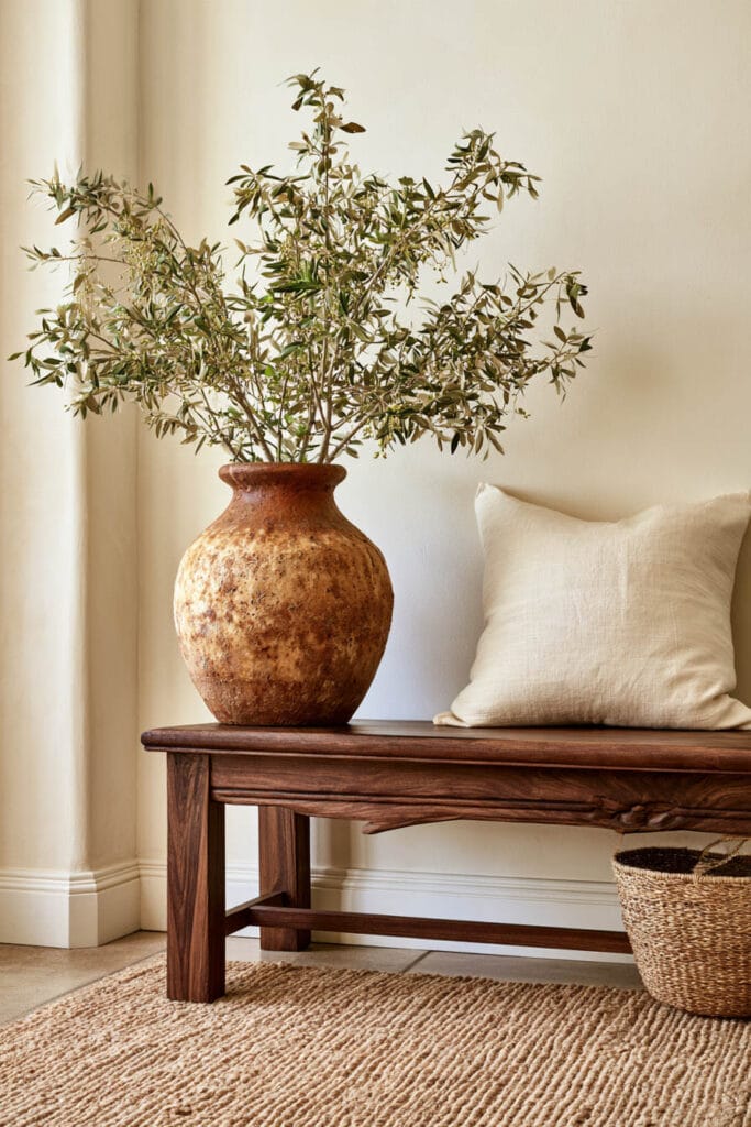 Afrohemian entryway with large weathered terracotta pottery vase, olive branches, dark wood bench, cream linen pillow, woven basket, and natural rug.