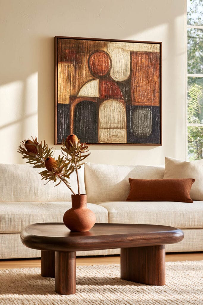 Afrohemian living room with large earth-toned figurative artwork, cream sofa, sculptural dark wood coffee table, terracotta vase, and textured rug.