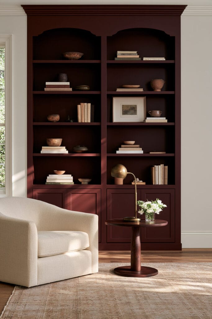 Deep burgundy built-in bookcases with curved decorative trim, cream rounded chair, wood side table, books, pottery, and brass lamp.