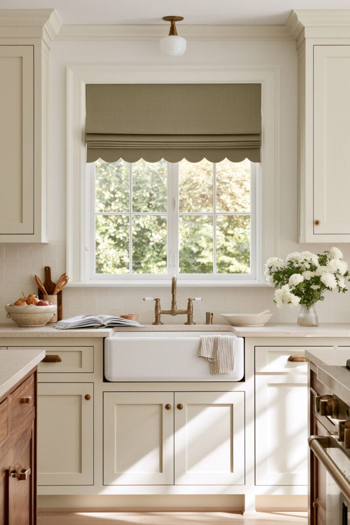 Bright cream kitchen with olive green scalloped Roman shade, farmhouse sink, warm wood accents, brass hardware, and fresh flowers.