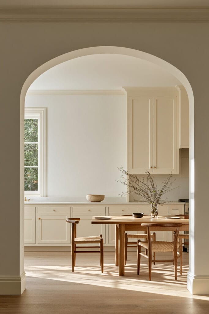Large arched doorway framing a warm neutral kitchen with natural wood dining table, woven chairs, and cream cabinetry.