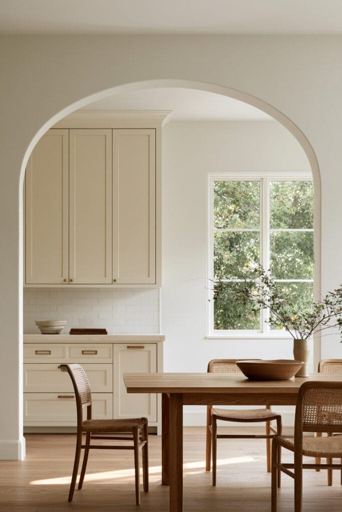 Large arched doorway connecting a warm neutral kitchen and dining room with wood furniture, cane chairs, and natural light.