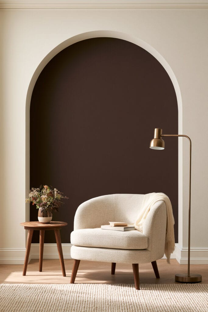 Chocolate brown painted arch framing a cozy reading nook with curved cream chair, round wood side table, and brass floor lamp.