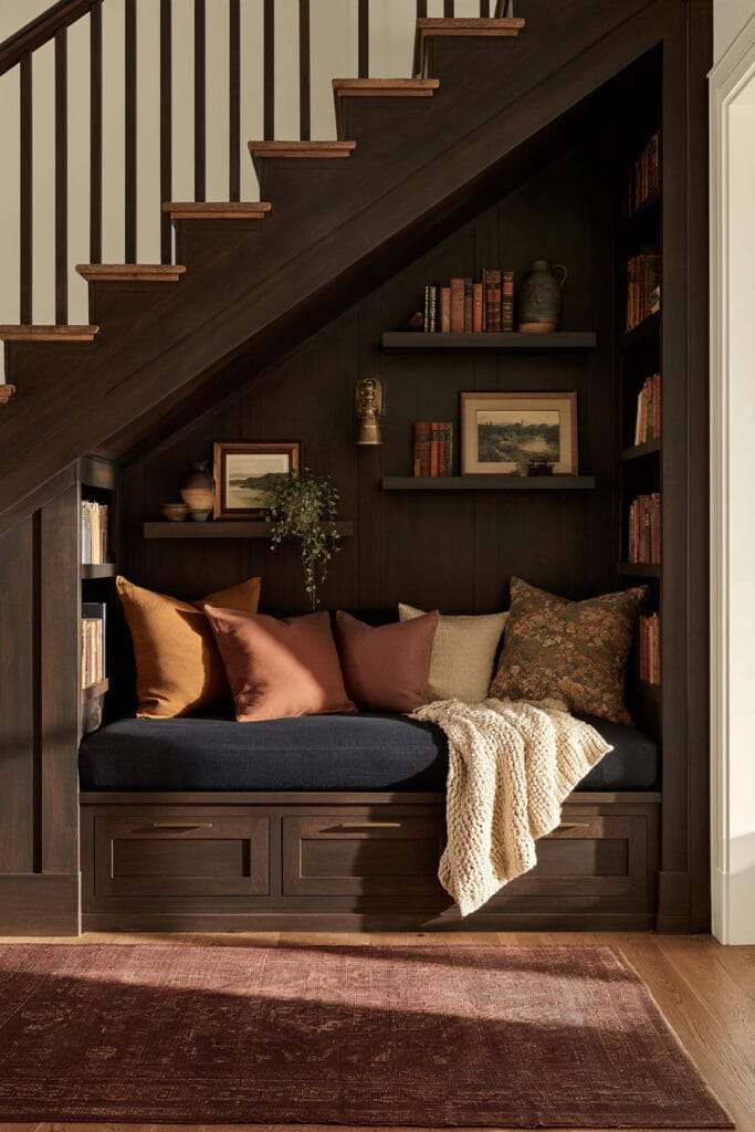Cozy under-stairs reading nook with dark wood paneling, built-in bench, charcoal cushion, rust pillows, bookshelves, storage drawers, and vintage burgundy rug.