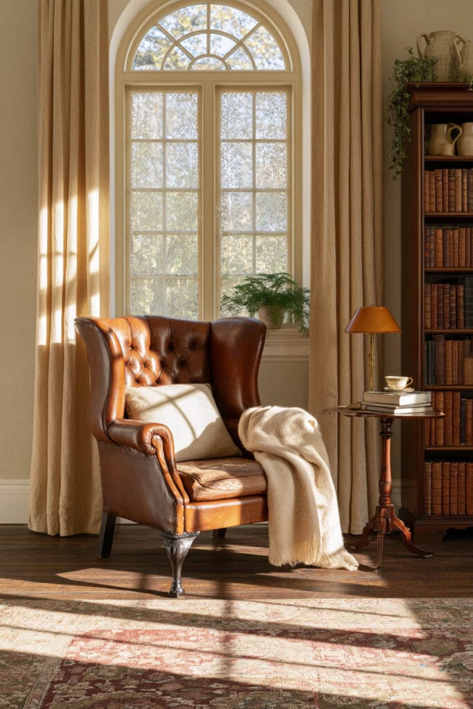 Cozy old-world reading nook with a cognac leather wingback chair, cream throw, arched window, linen curtains, antique wooden bookshelf, and vintage Persian-style rug.